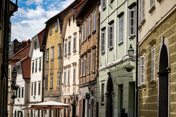Architectural view of historic townhouses with pastel-colored facades, traditional windows, and decorative elements lining a European street beneath summer clouds. Ljubljana, Slovenia. Gorni TRG