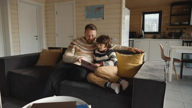 A white adult man and a school-aged boy sit together on a couch using a tablet in a wooden cabin living room. They wear sweaters and relax indoors, with a woman working in the kitchen background. - Powered by Adobe