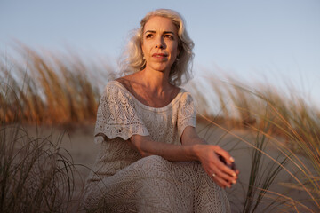 A serene image of a contemplative woman seated on sandy dunes. The warm sunset creates a peaceful...