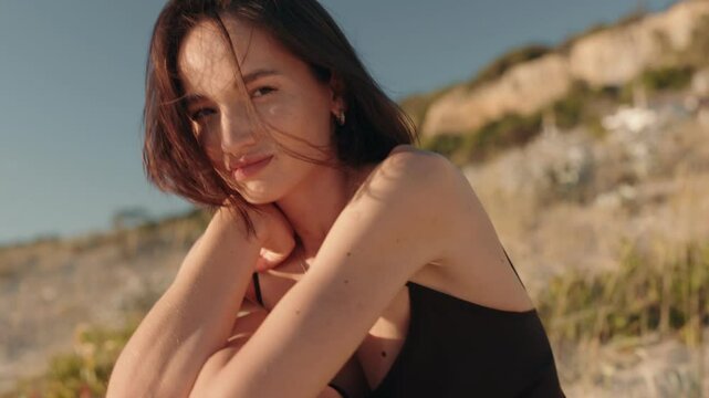 Attractive young woman sits peacefully on a sunny beach with the wind in her hair, enjoying the serene atmosphere and the warmth of the sun on her skin.