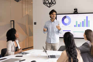 Young male professional presenting data to teammates standing in modern office next to large...