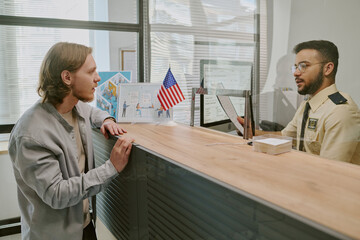 Person with light-colored hair talking to person in security uniform behind reception desk with...