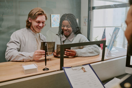 Smiling couple sitting at counter of public office, filling out forms and holding ID cards while engaging with official sitting behind glass partition