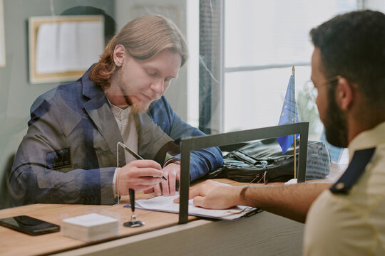 Individual with long hair signing documents in professional visa office environment while engaging with an officer across desk, showing attentive focus