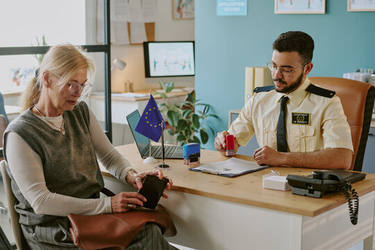 Senior woman sitting at immigration office with officer checking her documents. Desk features European Union flag, notepad, telephone, and device