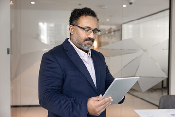 Middle-aged Indian analyst, businessman in formal suit standing in modern workspace office,...