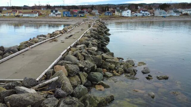 Drone footage of a quiet coastal village with an abandoned wharf reinforced by riprap along the calm St. Lawrence River in Baie‑des‑Sables, Quebec, 2025.