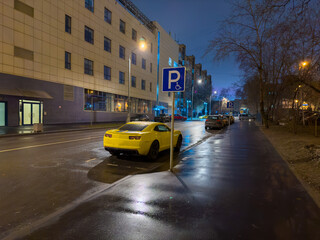 Fototapeta premium A vibrant yellow sport car is conspicuously parked on a wet city street at dusk, reflecting the glowing streetlights and the illuminated facade of a modern building. The low-angle shot captures the