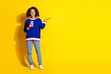 Smiling young woman in bright outfit holding phone and gesturing against vibrant yellow background showcasing trendy casual style