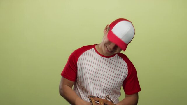 Young man in red baseball uniform and cap smiling over yellow background, holding glove and ball, radiating sporty enthusiasm.