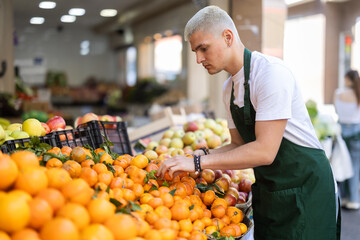 Young guy seller in apron puts fresh tangerines on display at vegetable market