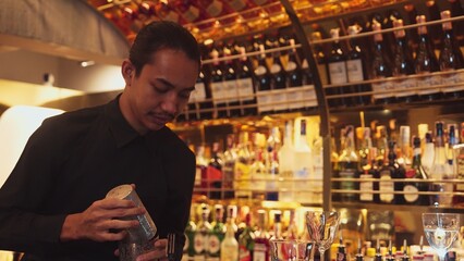 Smiling Asian smart bartender shaking with two hands in mixed cocktail in shaker preparing at luxurious counter bar, preparation special refreshment alcohol drink nightlife warm nightclub. Vinosity.