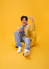 Happy young asian man sitting on bean bag eating popcorn and watching movie at the cinema home studio isolated on yellow background.