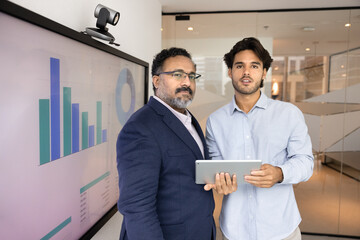 Young 30s Latin businessman holds tablet posing for photo with mature 45s Indian male colleague or company client standing together in office, monitor with infographic data nearby. Analytics, business