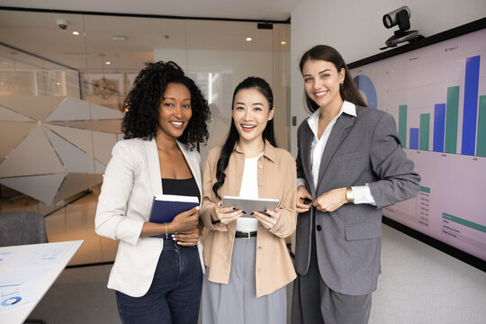 Three diverse professional women posing confidently in office meeting room, hold notebook and tablet, standing next to digital screen displays business graphs and analytics. Collaboration, leadership
