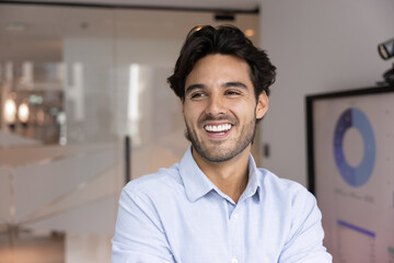 Fototapeta premium Cheerful young Hispanic businessman, looking away with smile, stands in modern workspace next to digital screen displaying business analytics bar and pie charts. Strategic vision, leadership, growth