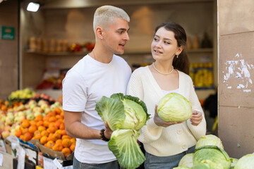 Young man and woman shopping in vegetable store and buy cabbage. They carefully examine goods,...