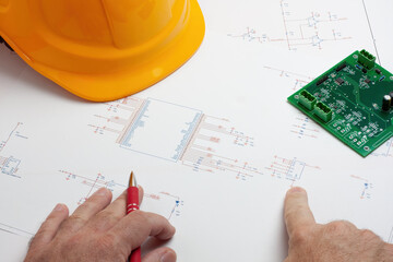 Hands working on electronic circuit schematic with PCB and hard hat
