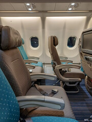 The interior of an airplane cabin is shown, featuring economy class seats. The seats are upholstered in a mix of brown and teal fabric, with adjustable headrests. A window is visible beside each seat