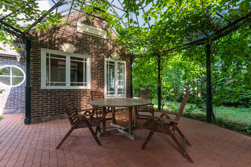 A beautiful ivy-covered gazebo with garden furniture, in the backyard of a brick house on a sunny...
