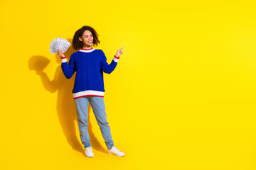 Smiling young woman in casual blue sweater holding money and pointing to the side on a vibrant yellow isolated background