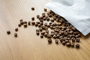 Medium roasted coffee beans spilled out of a white paper bag onto a wooden surface.