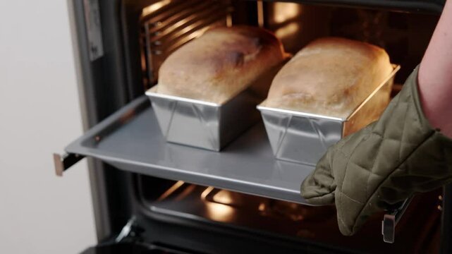 A person wearing an oven mitt removes two freshly baked bread loaves in metal pans from the oven, showcasing home baking.
