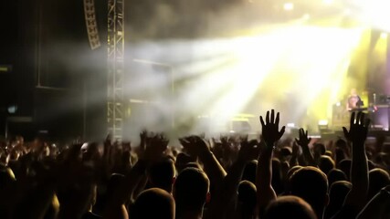 Crowd with raised hands enjoying live concert on large stage, bright lights and smoke create dynamic atmosphere, metal stage structures in background. Music festival atmosphere
