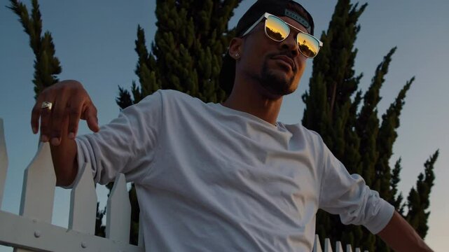 Homme portant des lunettes de soleil dor&eacute;es au coucher du soleil, attitude d&eacute;tendue devant une cl&ocirc;ture blanche
