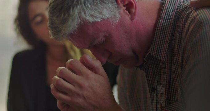 Man squeezes hands tightly and winces in visible pain during therapy session as group member offers comfort with gentle hand on shoulder in emotional moment