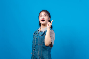 Excited young Asian woman in a denim dress pointing upward with a surprised and cheerful expression. Studio shot on blue background, ideal for creative advertising and promotions.