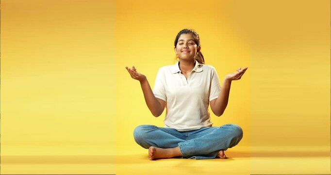 Girl Doing Mudra or Pranam, Indian Asian Schoolgirl Sitting Cross-Legged with Eyes Closed on Yellow Seamless Background Practicing Meditation with Hand Gesture for Calm Mindfulness and Inner Peace