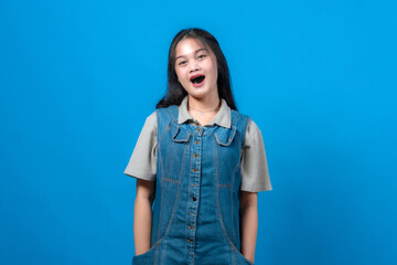 Fototapeta premium Surprised young Asian woman posing in front of a solid blue background, wearing a casual denim overall and short-sleeved shirt. She looks expressive, cheerful, and playful.
