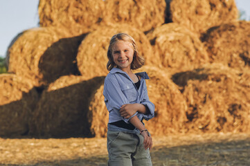 Horizontal portrait of smiling kid girl on the farm posing near rolls of hay bales, summertime in countryside, childhood and dreams