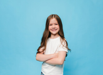 happy little brunette girl standing on blue background with arms crossed on chest