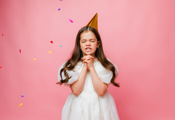Little girl in birthday cap rejoices and catches confetti on pink background