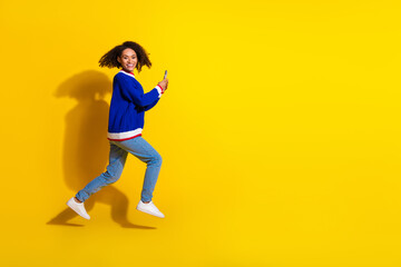Fototapeta na wymiar Cheerful young woman in a vibrant sweater jumps with energy and holds her phone against a bright yellow background