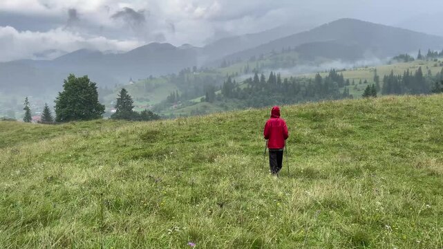 Travel woman in a red raincoat walking through scenic mountain hills