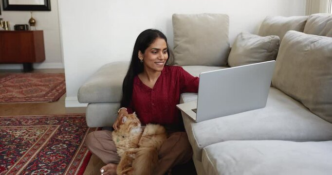 Positive relaxed adult Indian freelancer girl working in cozy comfortable home office, sitting on warm floor, petting furry cat, typing on laptop placed on soft couch, smiling