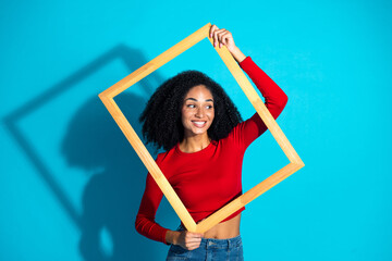 Young woman posing with a wooden frame in front of a bright blue background wearing a stylish red...