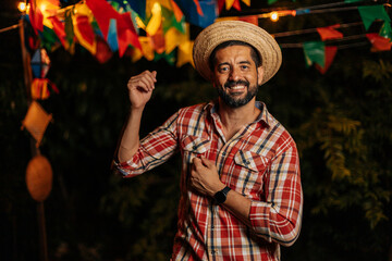 Happy man wearing a straw hat and plaid shirt at a traditional Brazilian festa junina. Colorful flags and warm lights decorate the festive night background