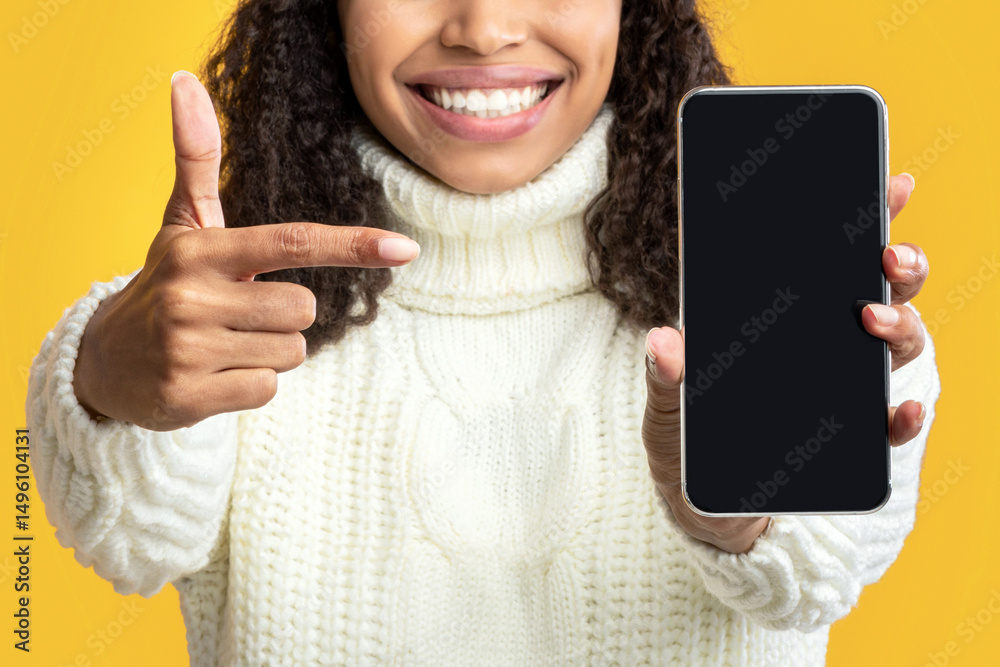 Poster Mobile Application. Black lady in winter hat holding and pointing at empty cellphone screen over orange studio background. Happy woman showing and indicating device display for mock up