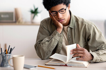 Bored African American teenager reading textbook, getting ready for complicated exam at home. Tired...