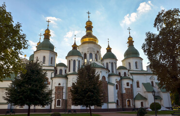 Majestic gold and green domes with crosses of a Kyiv St. Sophia Cathedral stand tall against a bright blue sky, framed by trees