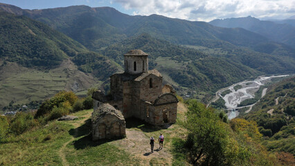 Fototapeta premium Panoramic aerial view of a very ancient dilapidated Christian temple on top of a mountain surrounded by mountains. There is a mountain river below. Shooting from a drone.