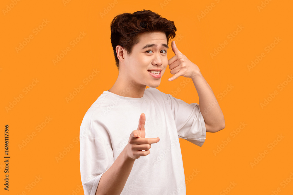 Poster playful asian man pointing index finger at camera, doing phone gesture saying call me back, talking 