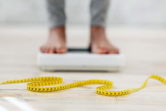 Unrecognizable black lady standing on scales, checking her weight, focus on measuring tape. Cropped view of African American woman fighting excess pounds, using slimming diet
