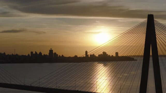 Drone flying between the cables of the San Roque Gonz&aacute;lez de Santa Cruz Bridge at sunset, with the skyline of Posadas in the background.