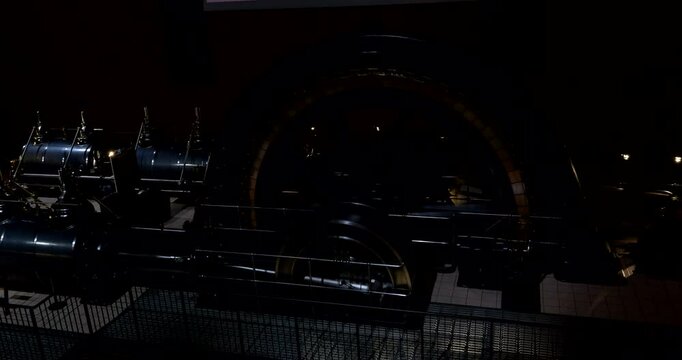 Large vintage generator with rotating central flywheel and mechanical components illuminated in dramatic red lighting, set in a tiled industrial hall with protective barriers