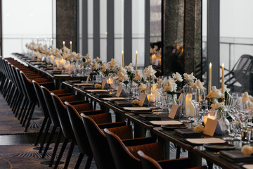  Panoramic view of a refined black table setting with white roses, handmade plates, natural wood chairs, and glasses. Elegant decor with fresh flowers for a sophisticated event atmosphere.